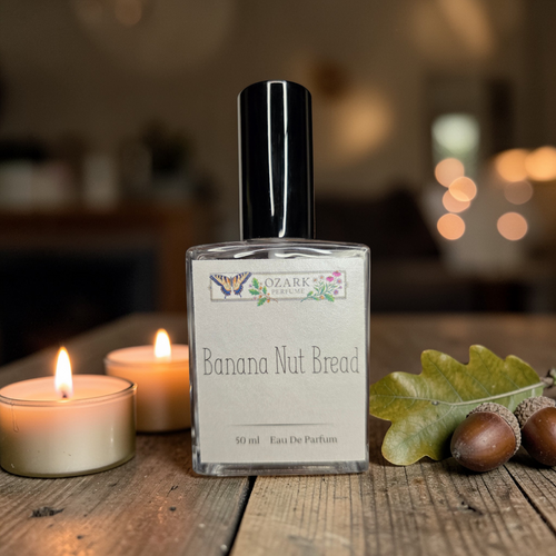 Perfume bottle labeled 'Banana Nut Bread' by Ozark Aromatics on a wooden surface with candles and leaves.