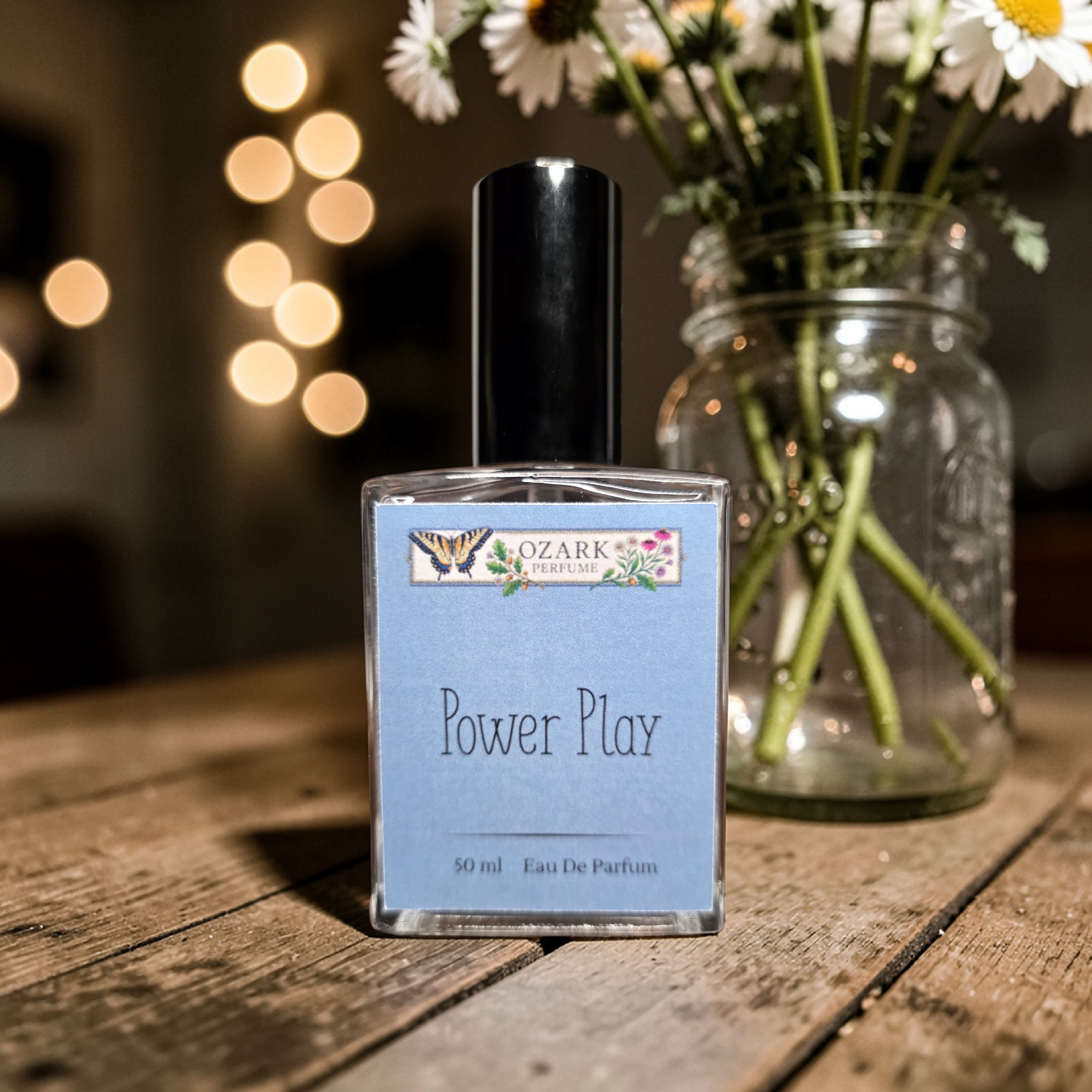 Perfume bottle labeled 'Power Play' by Ozark Perfume on a wooden surface with flowers in the background.