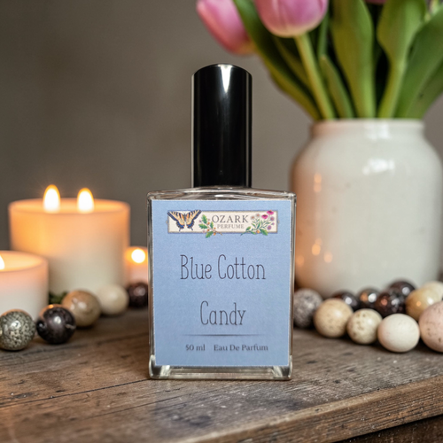 Perfume bottle labeled 'Blue Cotton Candy' by Ozark Perfume on a wooden surface with candles and flowers in the background.