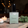 Perfume bottle labeled 'Element' by 'Ozark Perfume' on a wooden surface with candles and greenery.