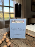 Perfume bottle labeled 'Vanilla Moderne' by Ozark Parfums on a wooden surface with a window in the background.