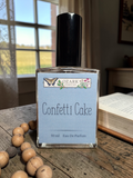 Perfume bottle labeled 'Confetti Cake' by Ozark Perfume Co. on a wooden surface with a window in the background.