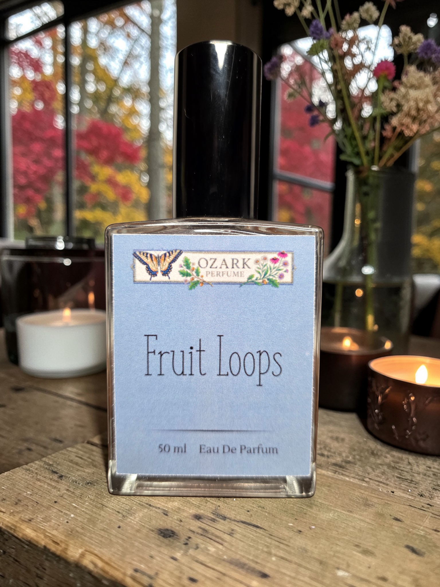 Eau de Parfum bottle labeled 'Fruit Loops' by Ozark Aromatics on a wooden surface with candles and flowers in the background.