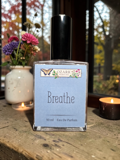 Ozark Essentials 'Breathe' eau de parfum bottle on a wooden surface with candles and flowers in the background.