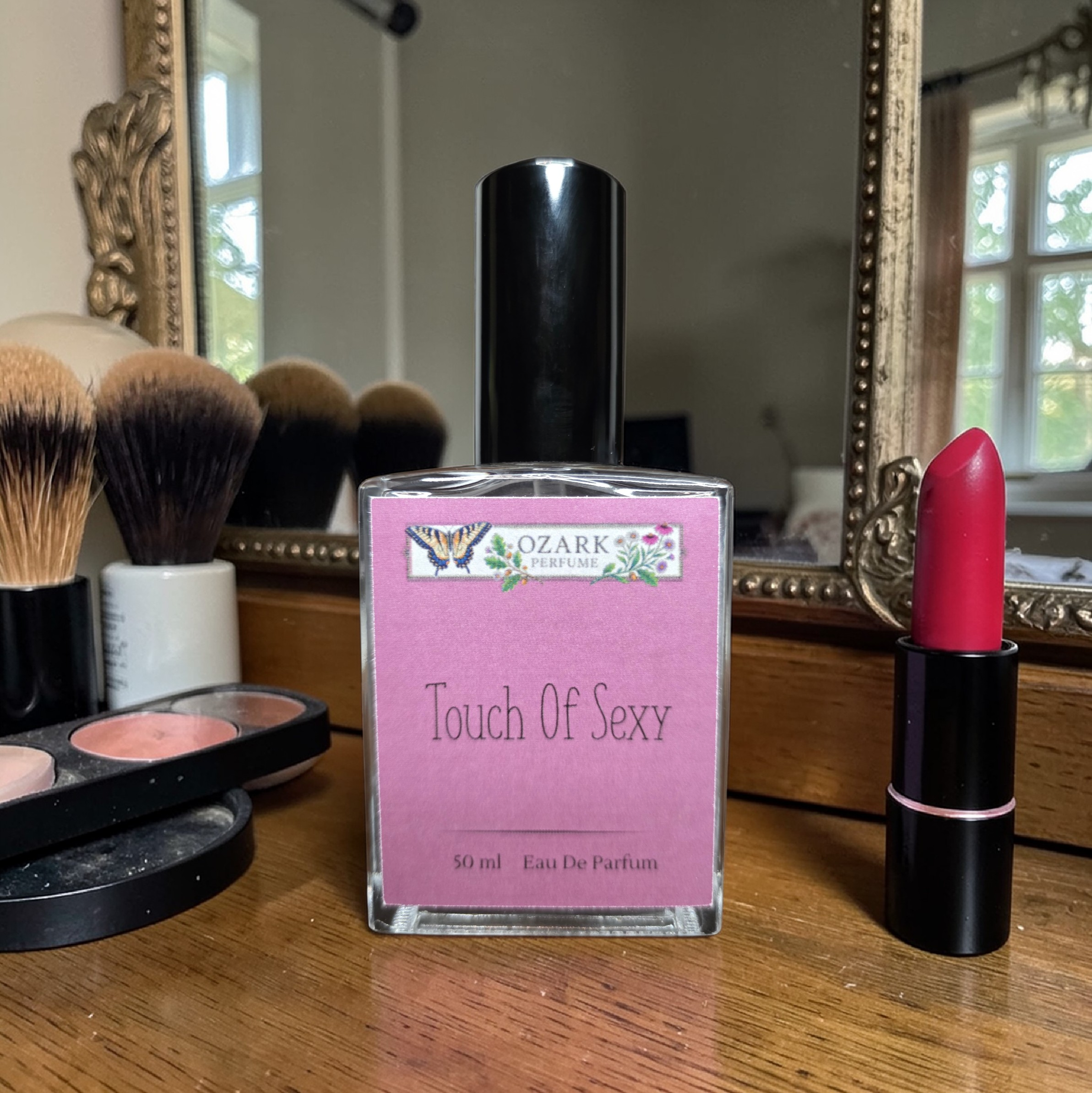 Perfume bottle labeled 'Touch Of Sexy' by Ozark Perfume, pink lipstick, and makeup brushes on a wooden surface.