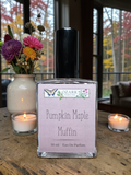 Bottle of Ozark Perfume Pumpkin Maple Muffin perfume on a wooden table with candles and flowers in the background.