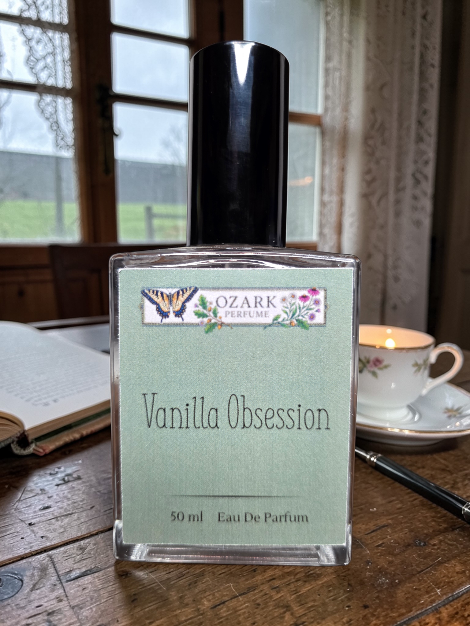 Perfume bottle labeled 'Vanilla Obsession' by Ozark Perfume on a wooden table with a book and tea cup in the background.
