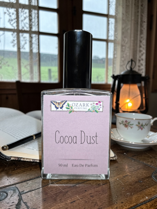 Perfume bottle labeled 'Cocoa Dust' by Ozark Aromatics on a wooden table with a scenic background.