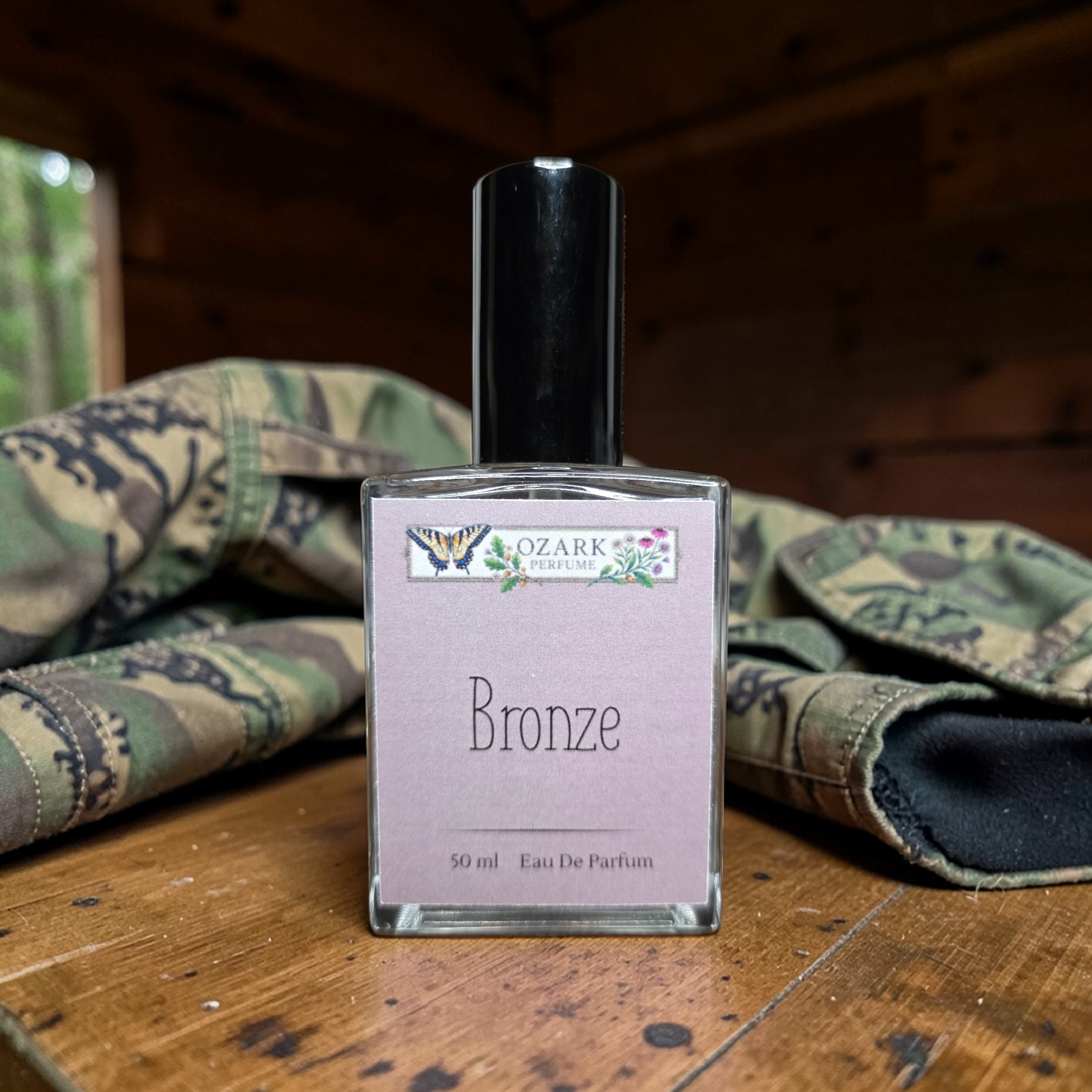 Perfume bottle labeled 'Bronze' by Ozark Perfume on a wooden surface with camouflage fabric in the background.