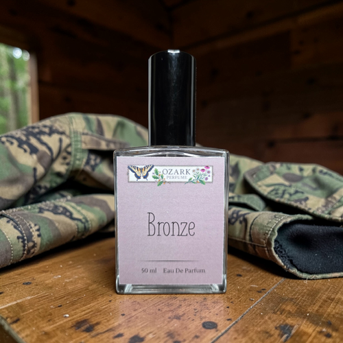 Perfume bottle labeled 'Bronze' by Ozark Perfume on a wooden surface with camouflage fabric in the background.