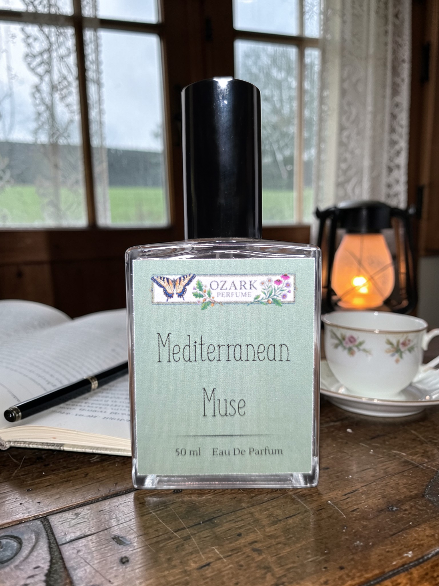Perfume bottle labeled 'Mediterranean Muse' by Ozark Trading Co on a wooden table with a cup and book in the background.