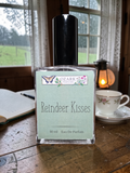 Perfume bottle labeled 'Reindeer Kisses' by Ozark Aromatics on a wooden table with a cup and book in the background.