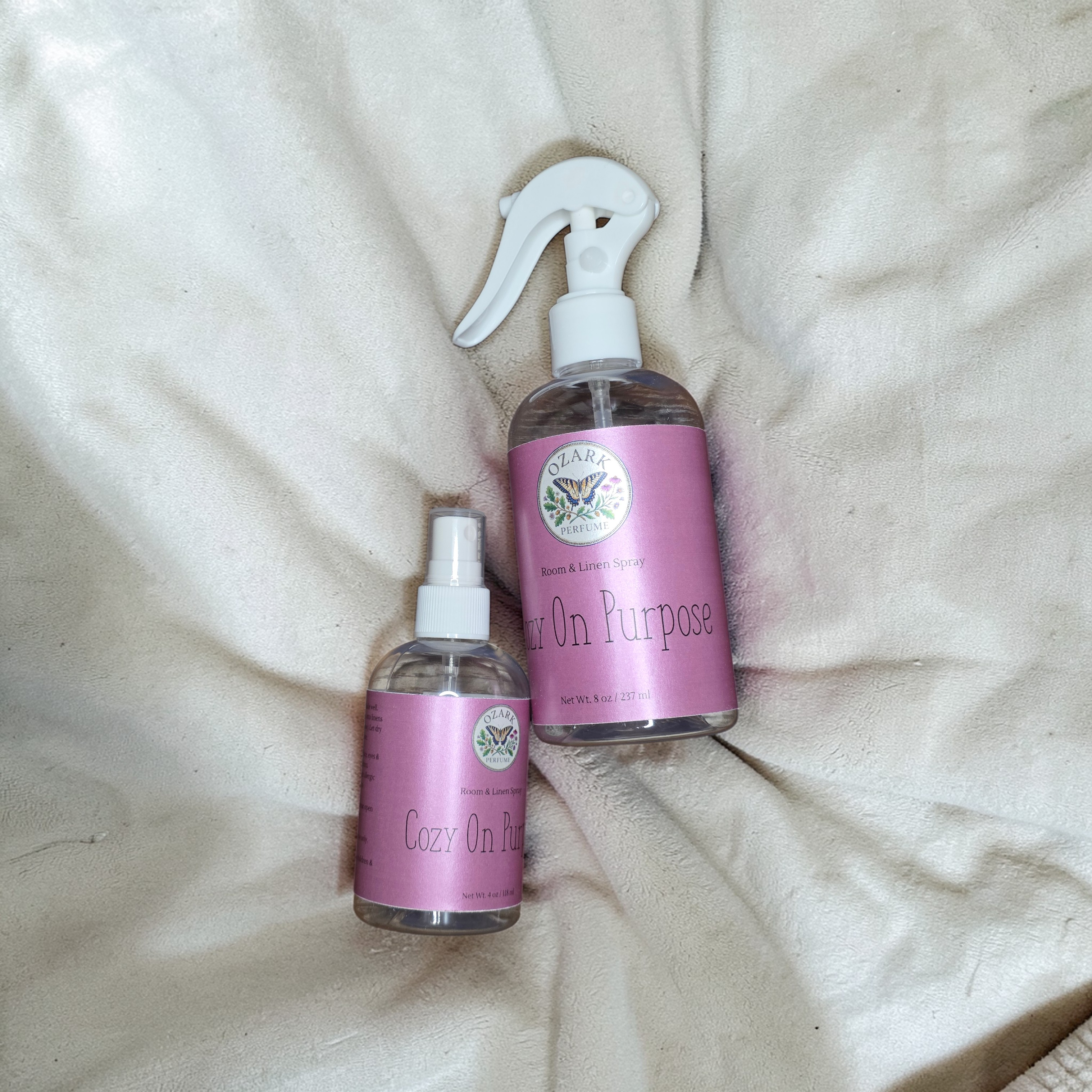 Two bottles of 'Cozy On Purpose' spray on a beige fabric background