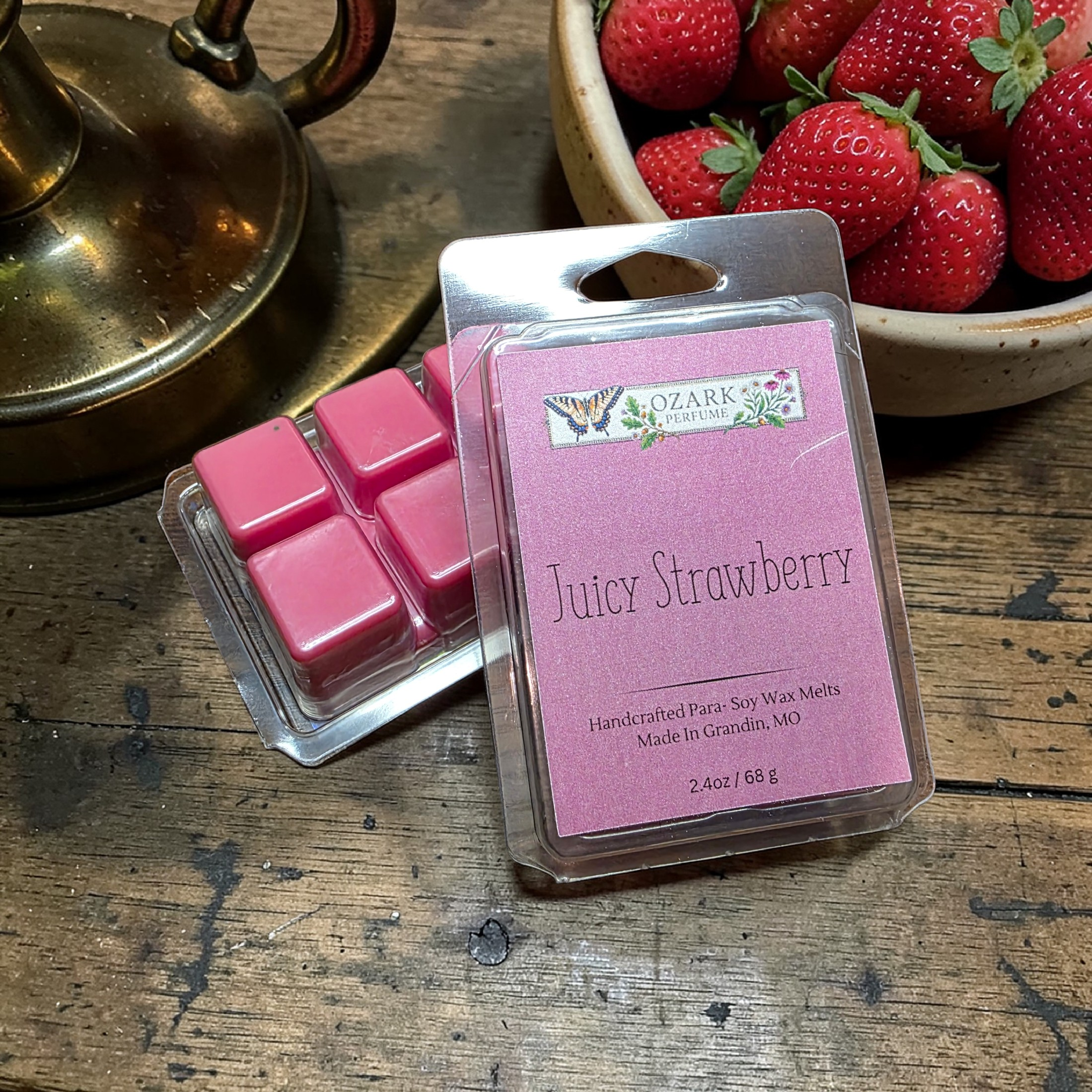 Pink wax melts in a package labeled 'Juicy Strawberry' on a wooden surface with strawberries and a brass lamp.