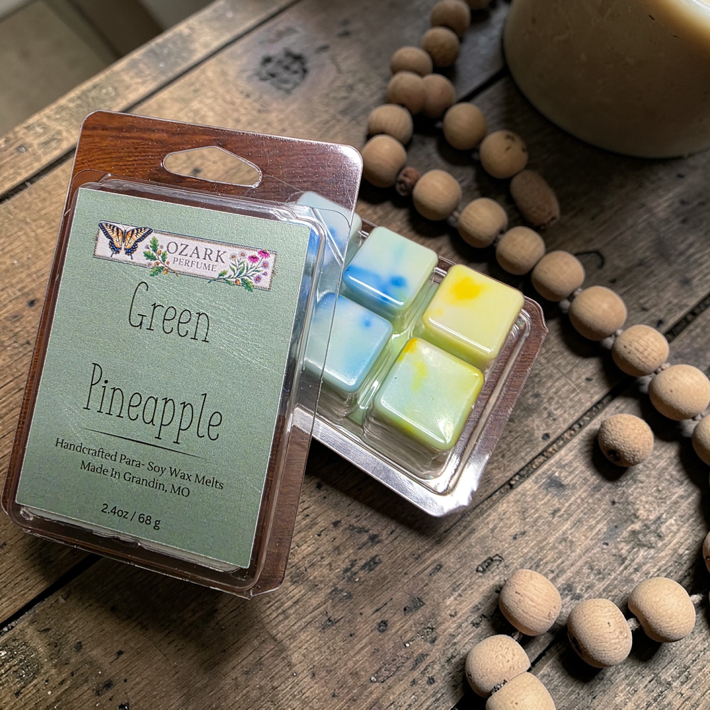Packaged wax melts labeled 'Green Pineapple' by Ozark Aromas on a wooden surface with beads.