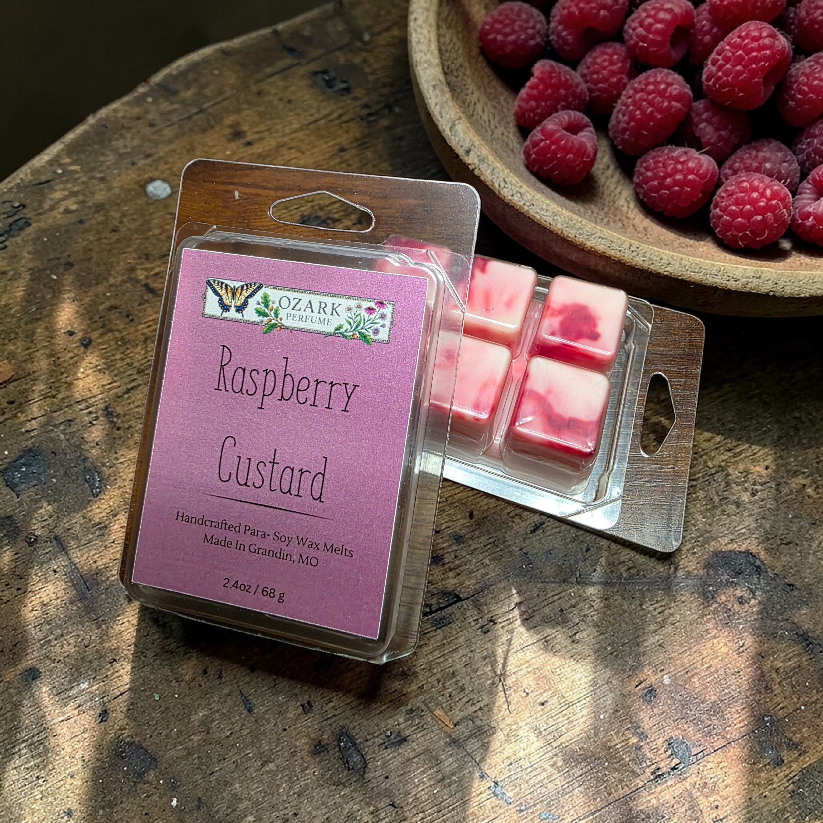 Packaged raspberry custard wax melts with a bowl of raspberries on a wooden surface