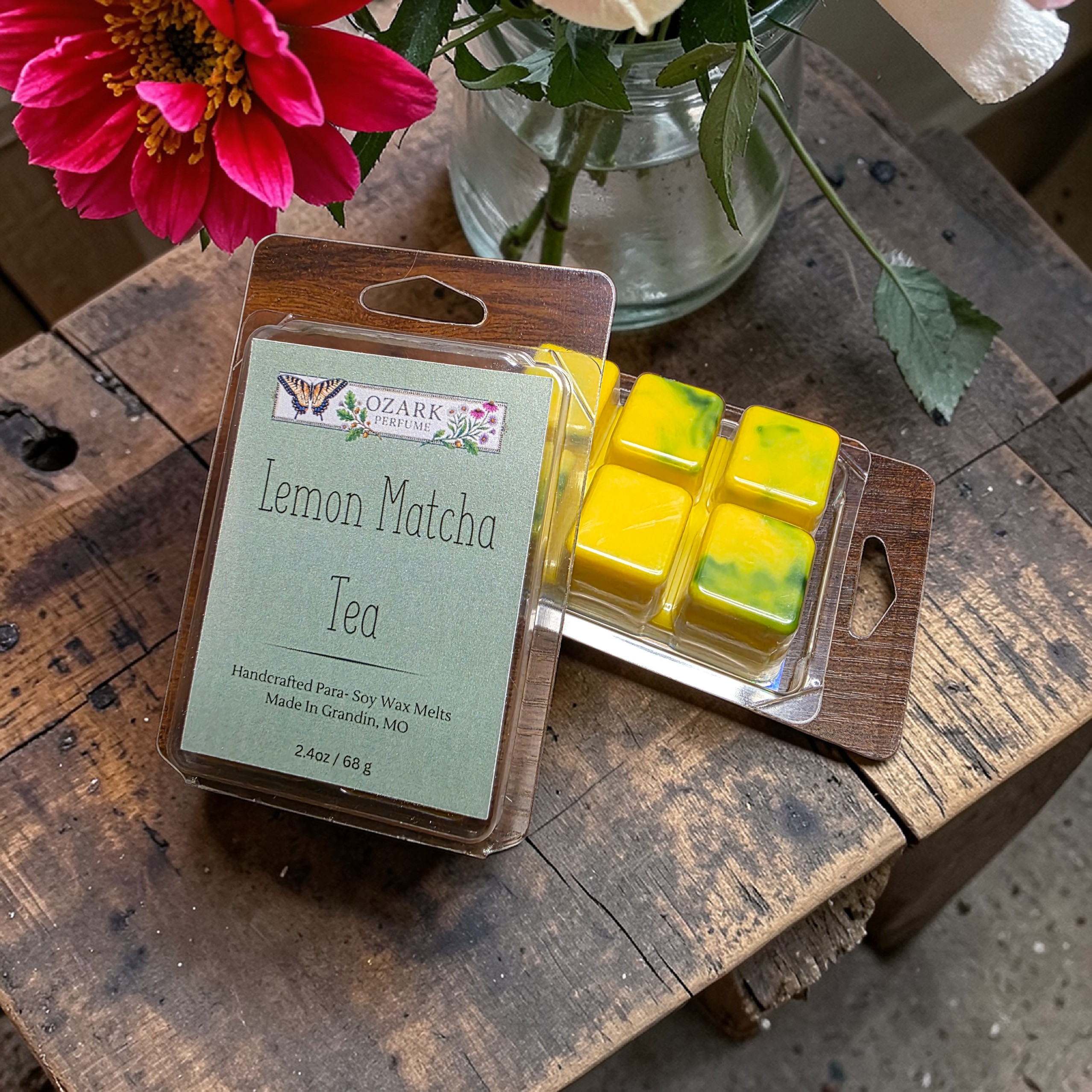Packaged lemon matcha tea wax melts on a wooden surface with a pink flower in the background.
