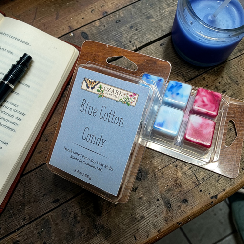 Packaged wax melts labeled 'Blue Cotton Candy' by Ozark Aromatics on a wooden surface with a candle and notebook.
