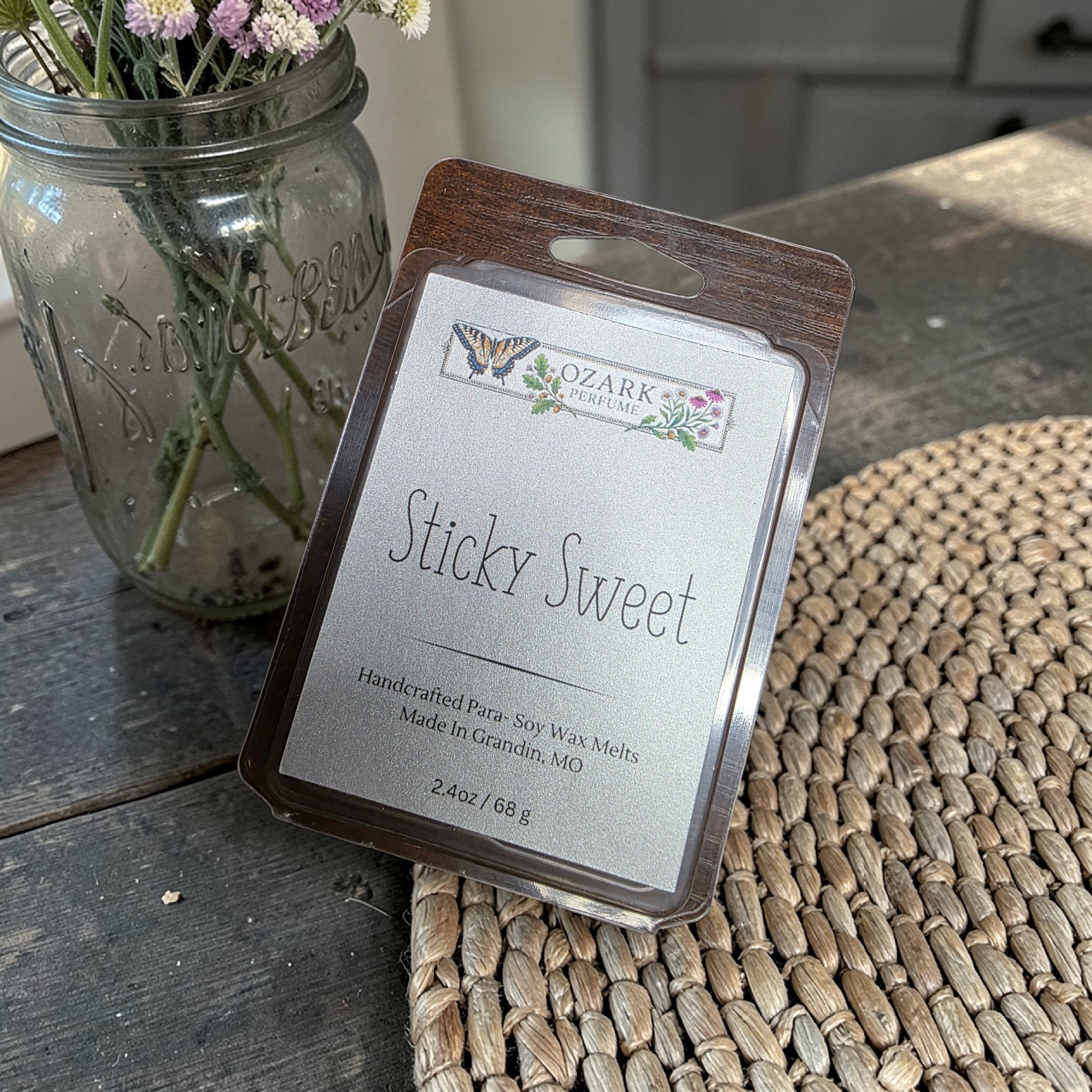 Ozark Trail wax melt labeled 'Sticky Sweet' on a woven mat with a jar of flowers in the background.