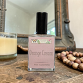 Perfume bottle labeled 'Orange Cream' by Ozark Perfume on a wooden surface with a candle and beads in the background.