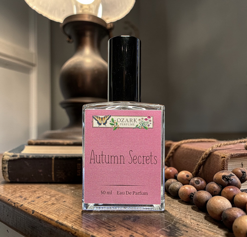 Perfume bottle labeled 'Autumn Secrets' by Ozark Aromatics on a wooden surface with books and berries in the background.