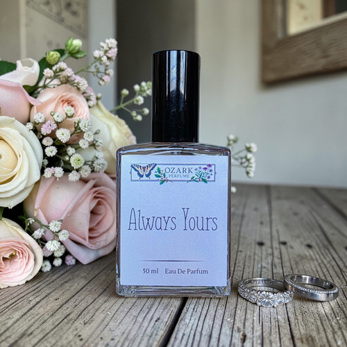 Perfume bottle labeled 'Always Yours' by Ozark Perfume with flowers and rings on a wooden surface.