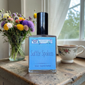 Perfume bottle labeled 'Softly Spoken' by Ozark Perfume on a wooden surface with flowers and a mug in the background.