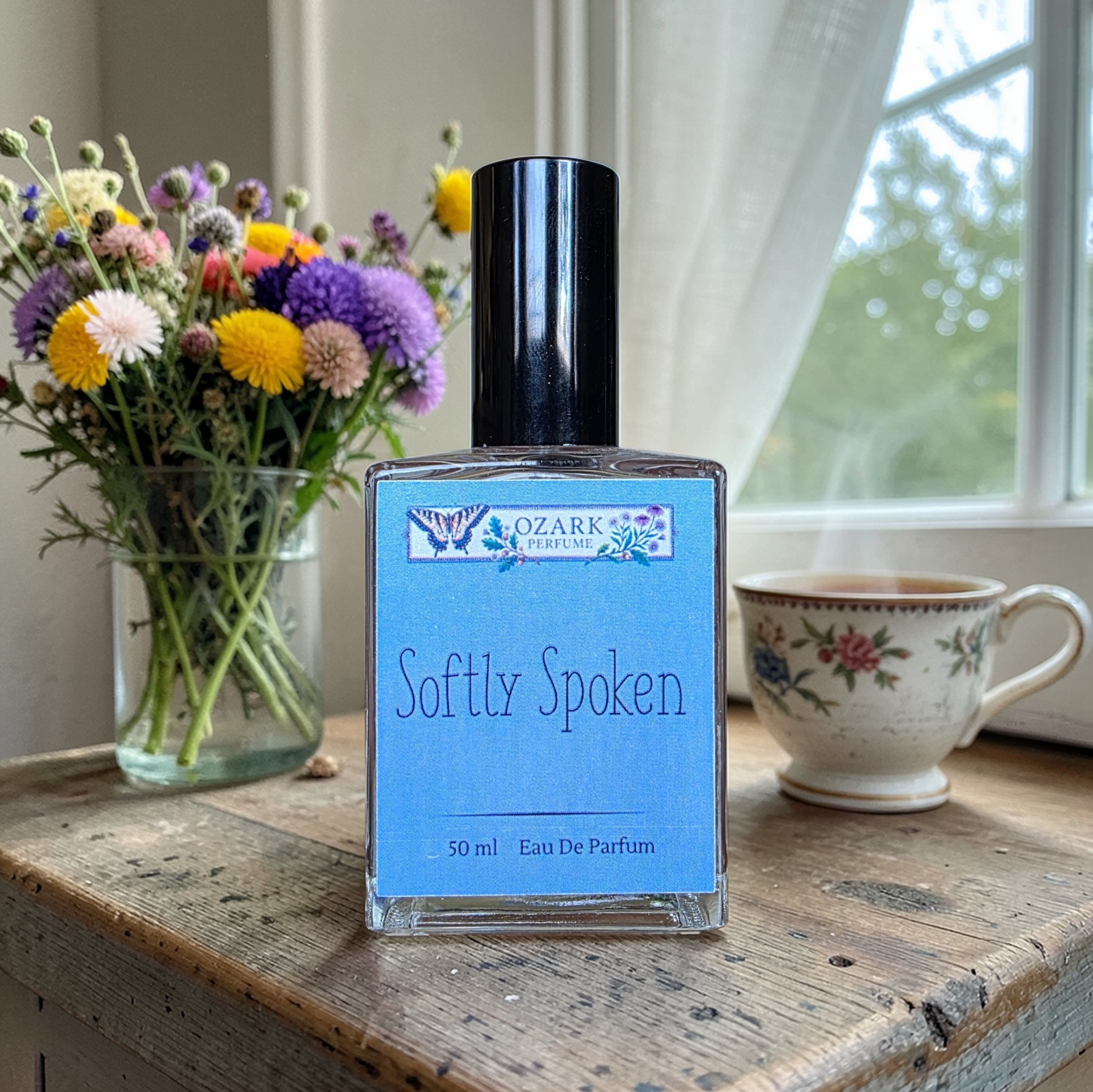 Perfume bottle labeled 'Softly Spoken' by Ozark Perfume on a wooden surface with flowers and a mug in the background.