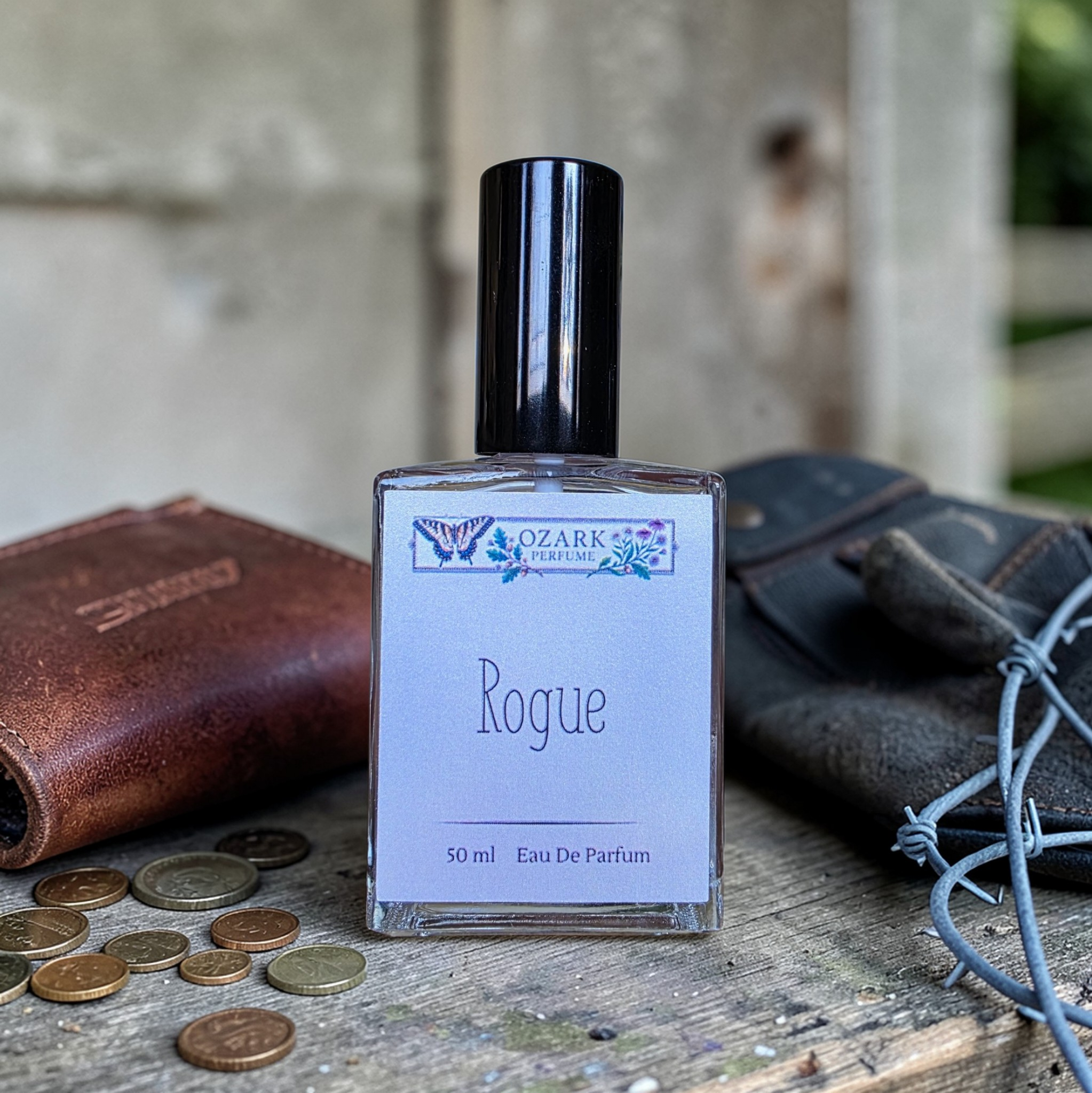 Perfume bottle labeled 'Rogue' by Ozark Parfum with coins and leather items on a rustic surface.