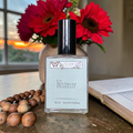 Perfume bottle labeled 'Whimsy' by Ozark Aromatics on a table with flowers and beads.