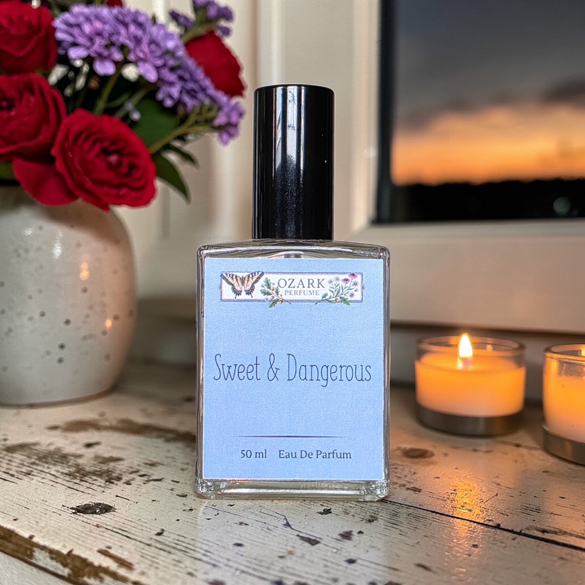 Perfume bottle labeled 'Sweet & Dangerous' by Ozark Aromatics on a wooden surface with flowers and candles in the background.