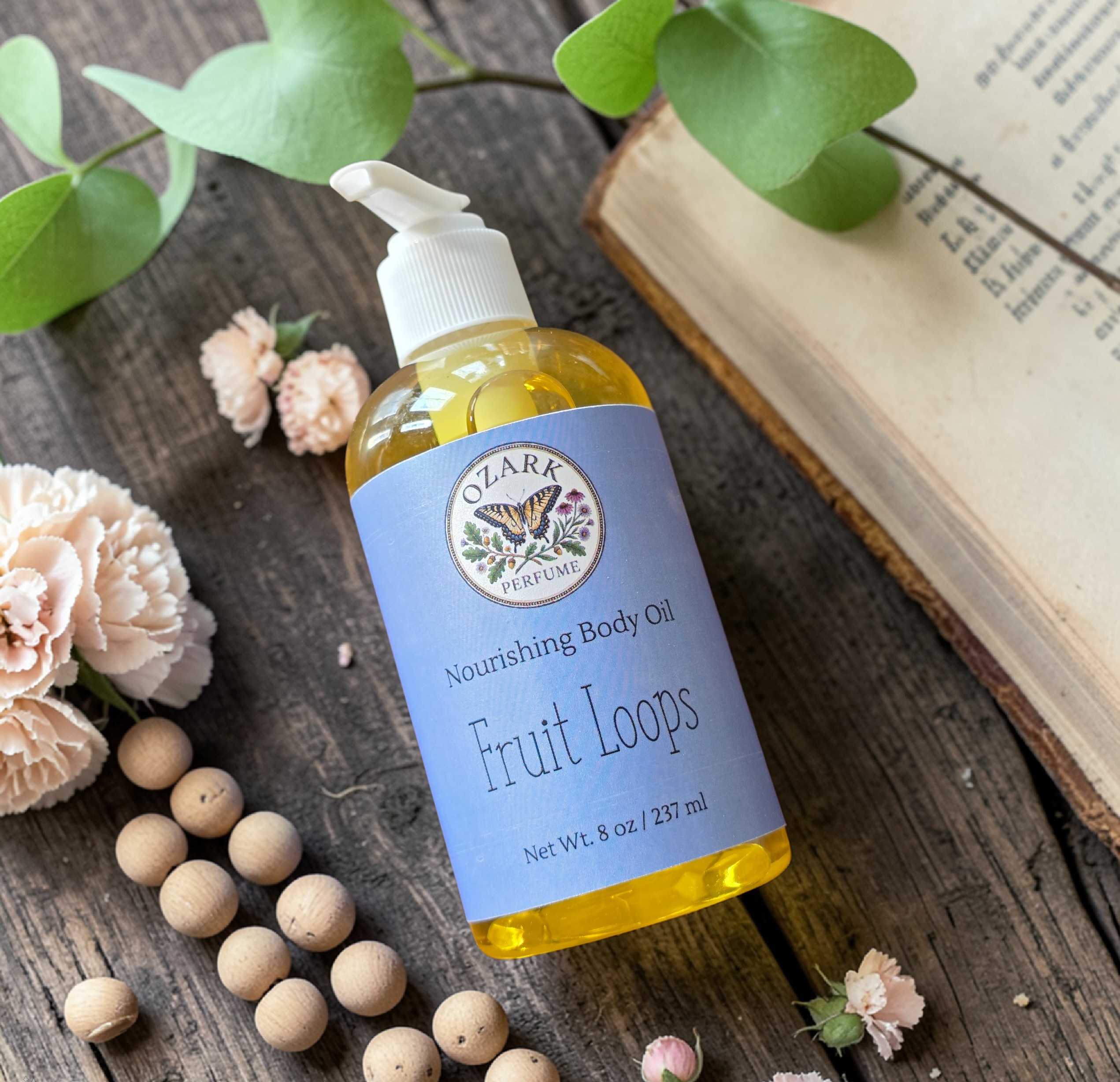 Bottle of Nourishing Body Oil labeled 'Fruit Loops' on a wooden surface with flowers and an open book.