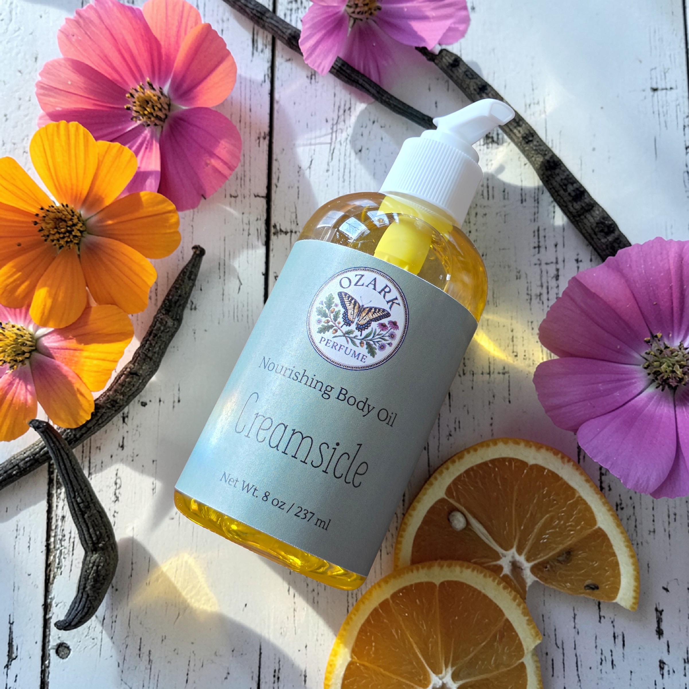 Body oil bottle labeled 'Creamsicle' with flowers and vanilla beans on a wooden surface
