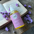 Bottle of 'Sugar Puff' body oil on an open book with purple flowers