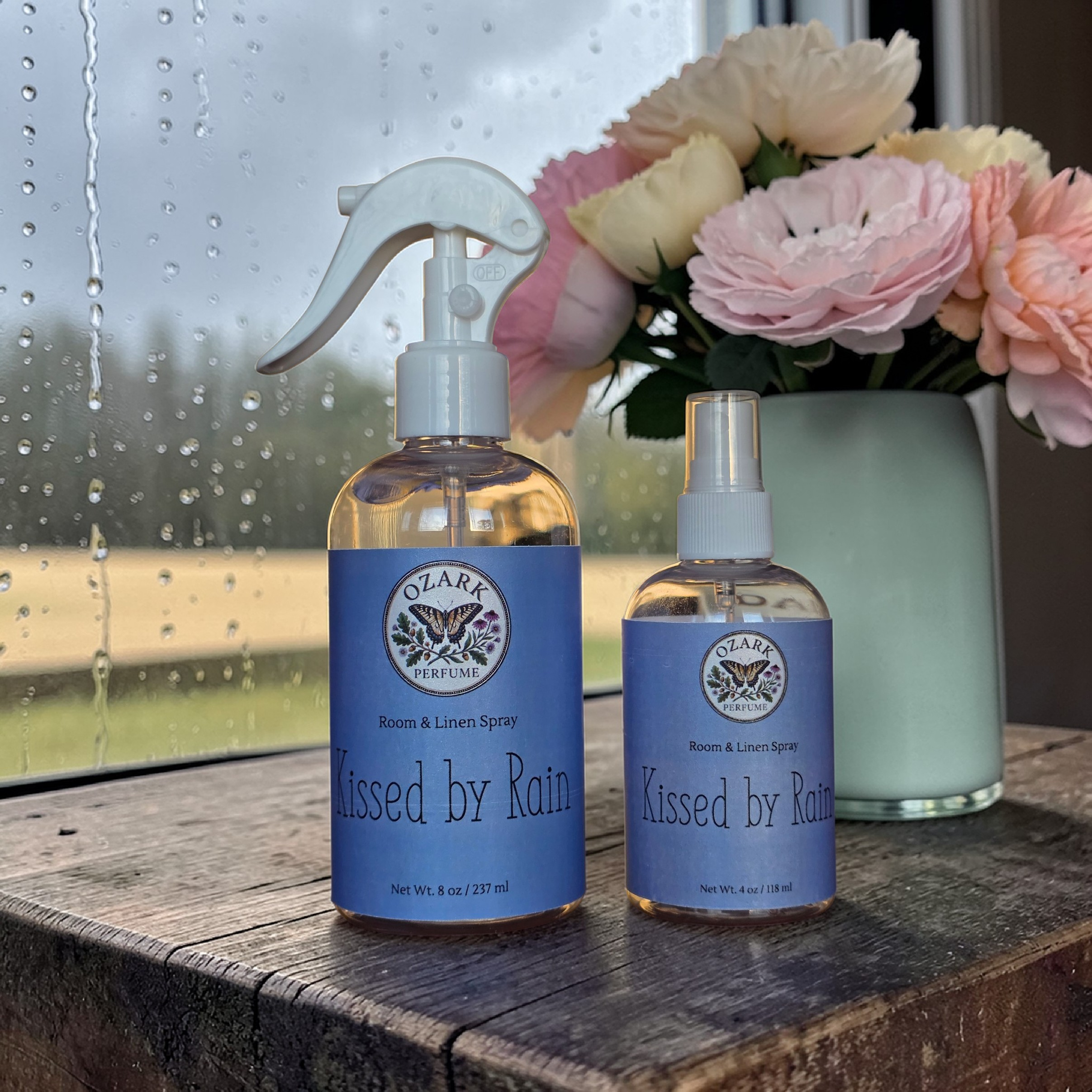 Two bottles of Ozark Perfume 'Kissed by Rain' on a wooden surface with flowers in the background.
