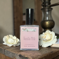Perfume bottle labeled 'Vanilla Milk' by 'Ozark Perfume' on a wooden surface with white flowers.