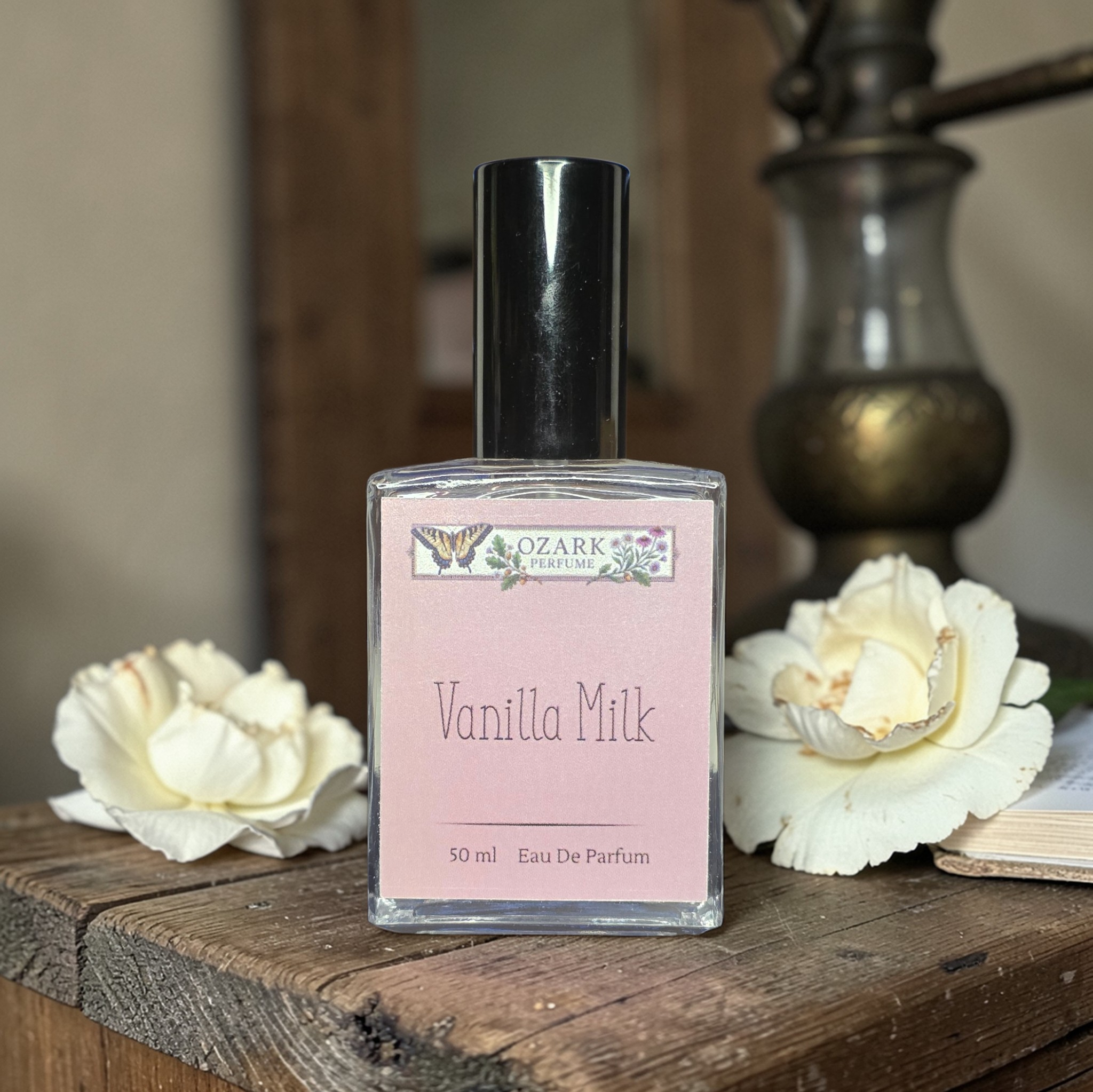 Perfume bottle labeled 'Vanilla Milk' by 'Ozark Perfume' on a wooden surface with white flowers.