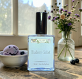 Perfume bottle labeled 'Blueberry Sorbet' on a wooden table with a bowl of bath bombs and a vase of flowers.