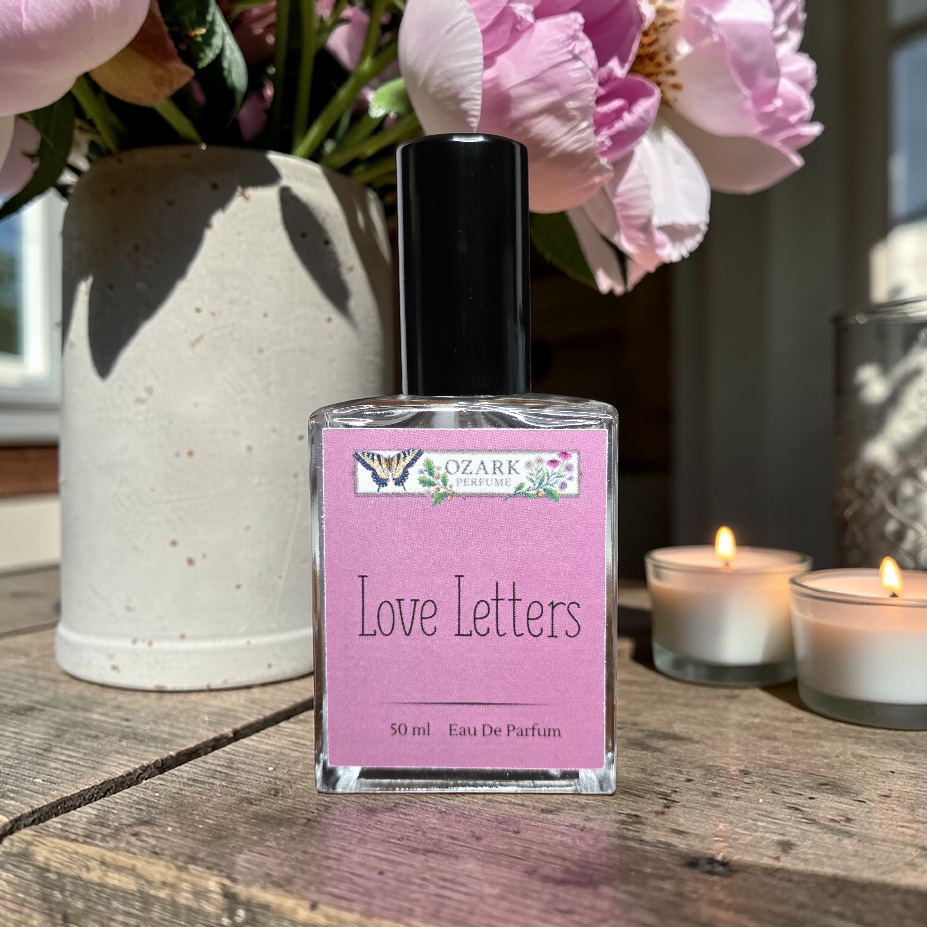 Perfume bottle labeled 'Love Letters' by Ozark with pink flowers and candles in the background.