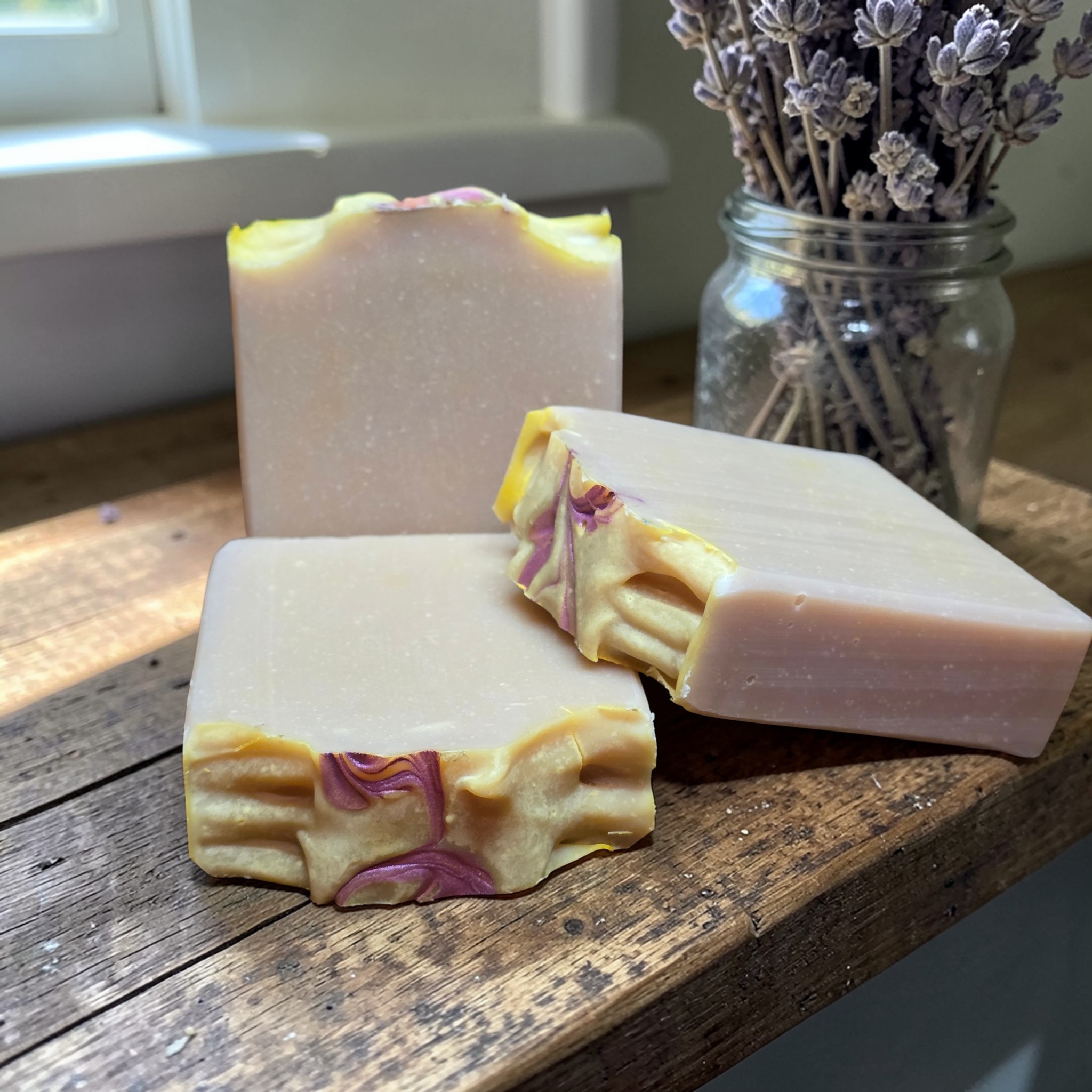 Three bars of soap with floral designs on a wooden surface, with a jar of lavender flowers in the background.