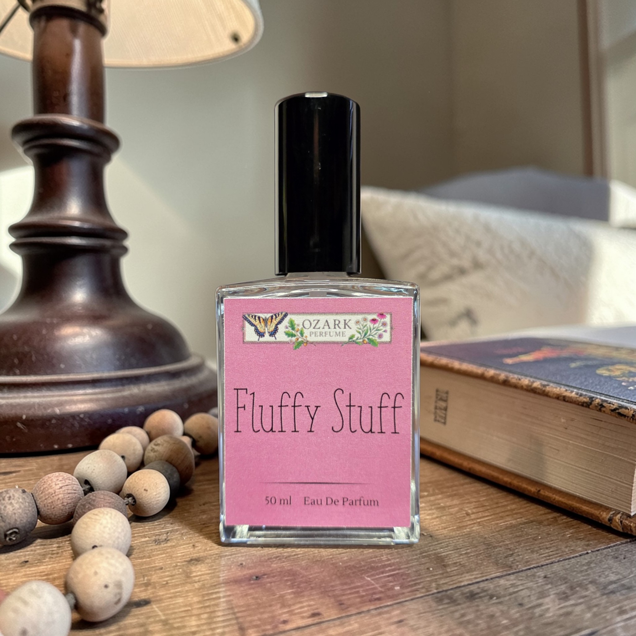 Perfume bottle labeled 'Fluffy Stuff' by Ozark Aromatics on a wooden surface with a lamp and book in the background.