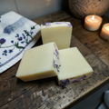 Three bars os soap with plavender buds on a wooden surface, with candles and a cloth in the background.