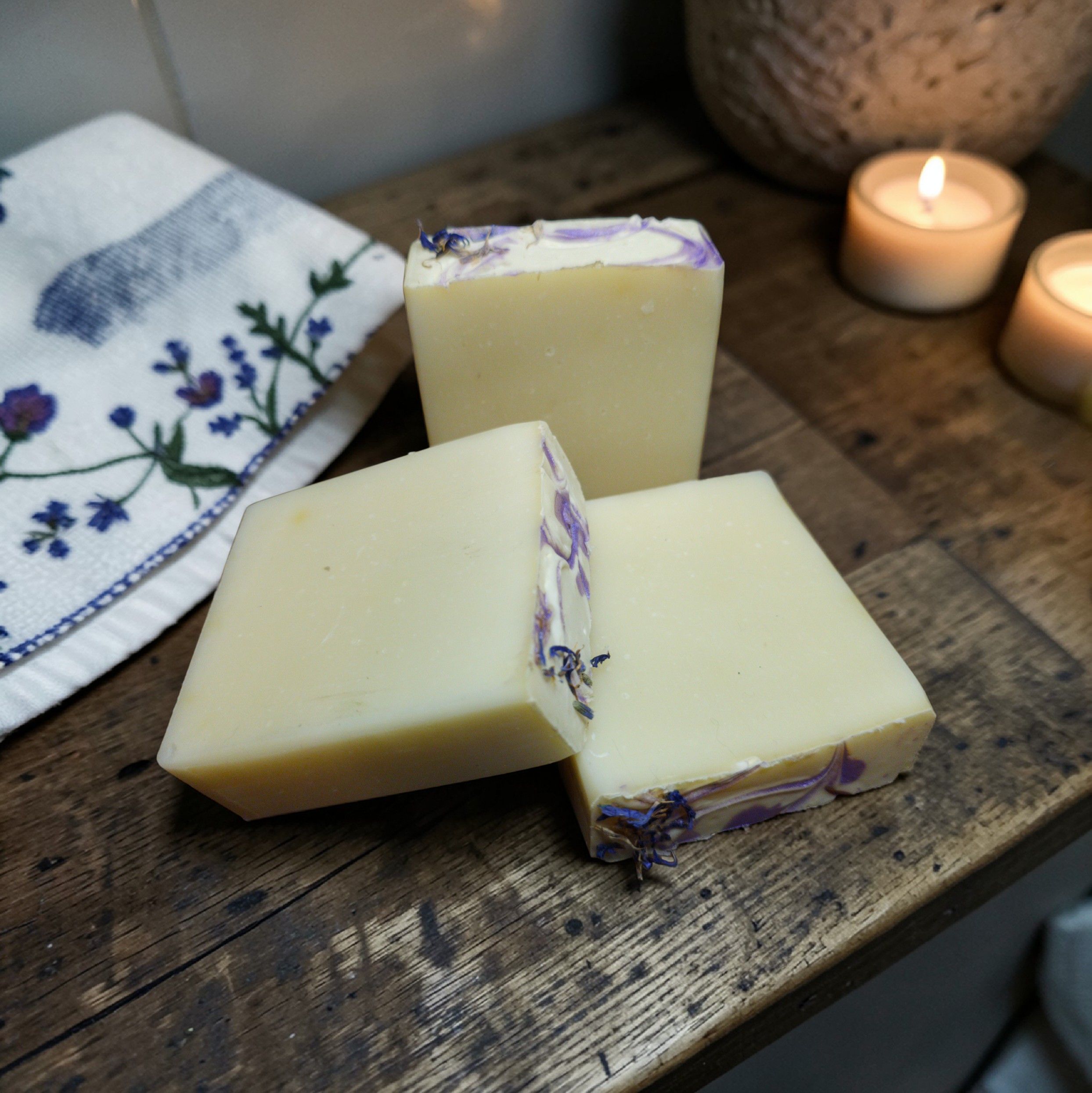 Three bars os soap with plavender buds on a wooden surface, with candles and a cloth in the background.