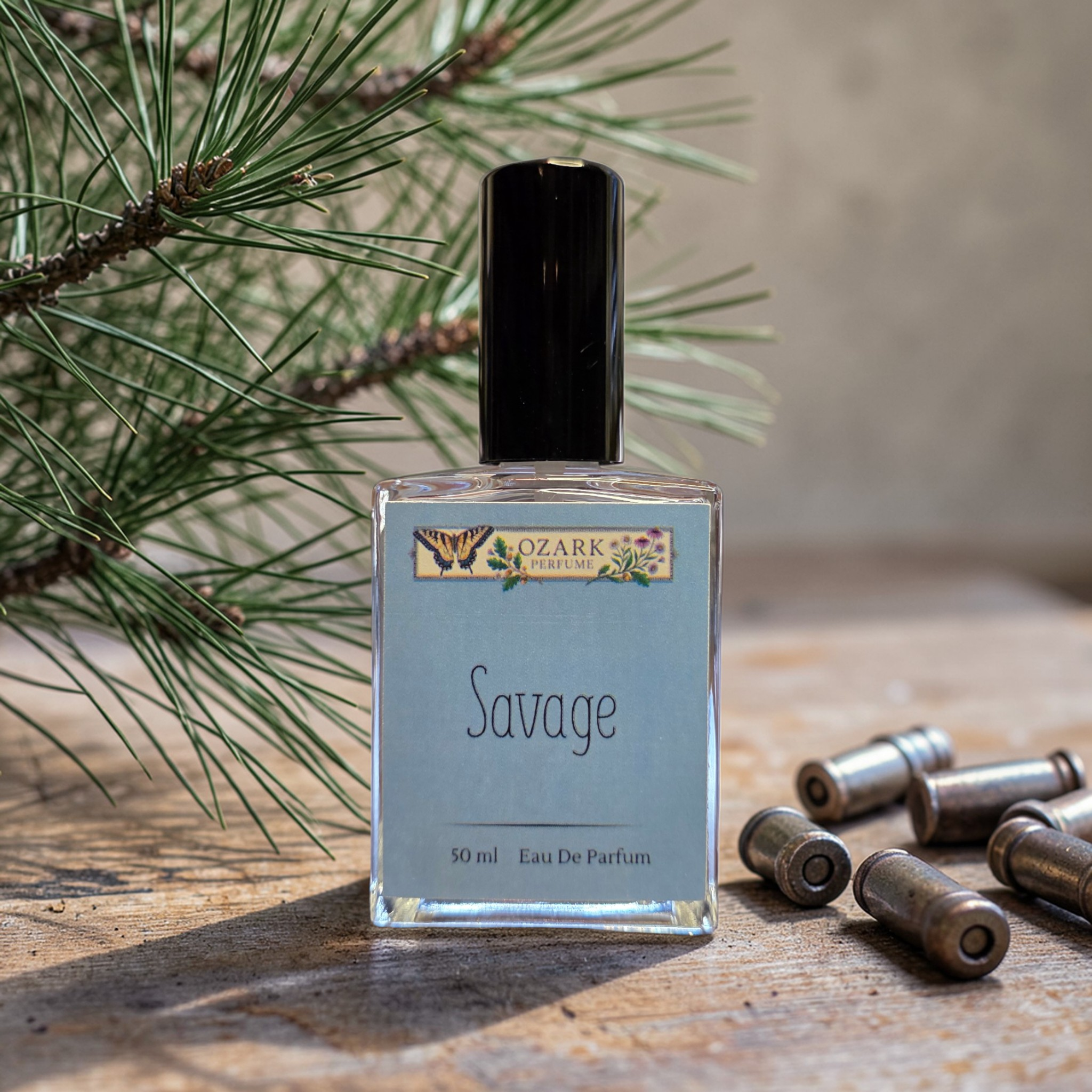 Perfume bottle labeled 'Savage' by 'Ozark' with pine branches and bullet casings on a wooden surface.